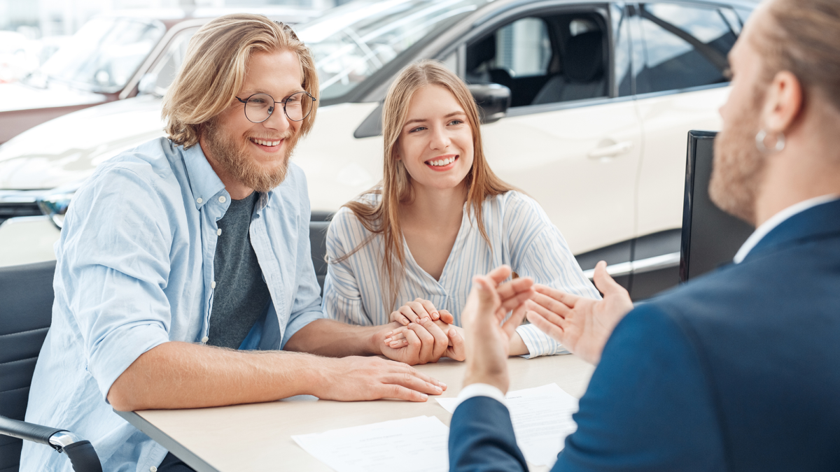 man and woman sitting at a table smiling and talking to someone in blue suite across the table from them and a white car in the background and papers on the table