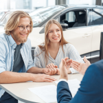 man and woman sitting at a table smiling and talking to someone in blue suite across the table from them and a white car in the background and papers on the table