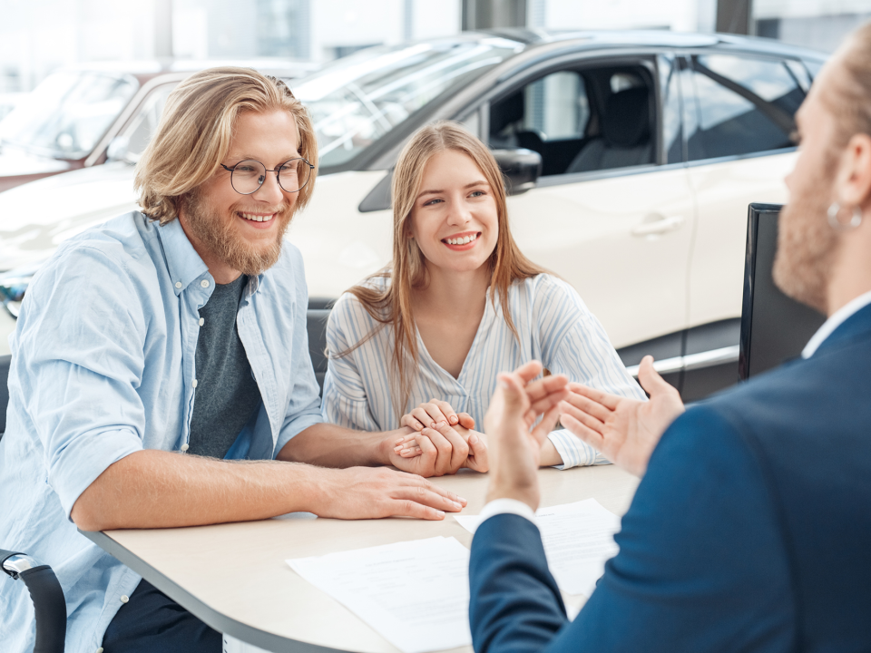 man and woman sitting at a table smiling and talking to someone in blue suite across the table from them and a white car in the background and papers on the table